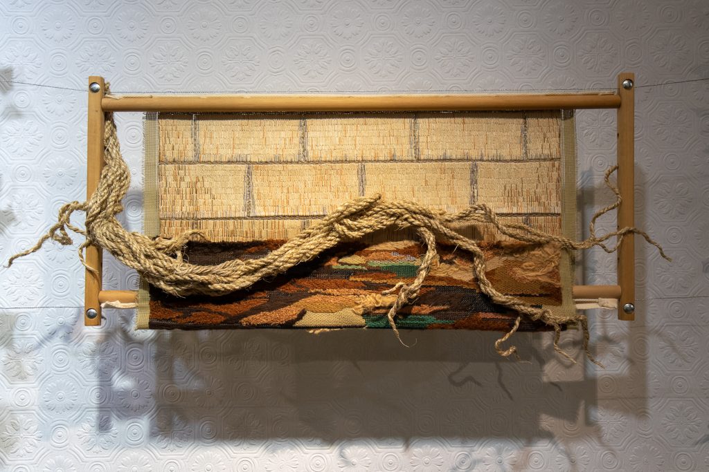 Embroidery of yellow bricks in a wooden rotating tapestry frame. In the foreground, there is a tick rope shaped and untwined resembling tree roots.